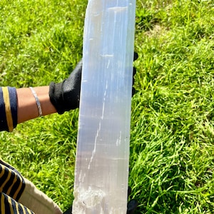 Può includere: Una grande torre di cristallo di selenite allungata con un aspetto bianco latte traslucido e distinte striature verticali. Il campione minerale presenta una struttura colonnare liscia con una consistenza cristallina ruvida alla base.