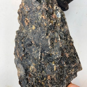 Puede incluir: Una roca grande, de color verde oscuro y negro, con una superficie rugosa y texturizada. La roca tiene una variedad de pequeñas inclusiones de color claro.
