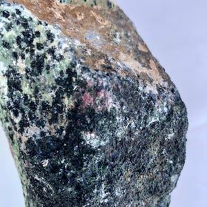 Könnte beinhalten: Nahaufnahme eines rauen, natürlichen Steins mit einer Vielzahl von Farben und Texturen. Der Stein weist Bereiche in Dunkelgrün, Schwarz, Braun und Rosa auf. Die Oberfläche ist uneben und scheint ein Mineralexemplar zu sein.