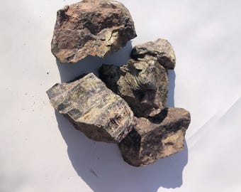 May include: A collection of rough, irregularly shaped rocks in shades of brown, gray, and green. The rocks have a textured surface and are arranged on a white background. The image is well-lit, highlighting the natural colors and details of the stones.
