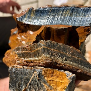 May include: Close-up of raw, uncut tiger's eye stone. The stone displays bands of blue, gold, and brown hues. The rough texture and natural patterns are visible. The stone is held in a hand.