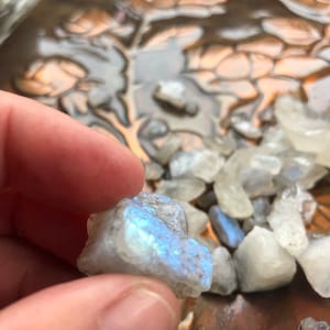 May include: A close-up of a small, white, iridescent gemstone with a blue sheen. The gemstone is being held in a hand, and there are other similar gemstones in the background.