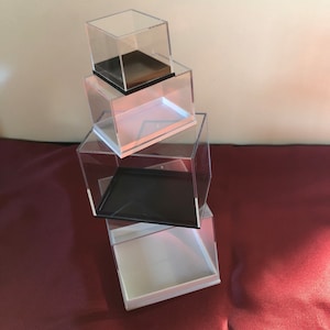 May include: Four clear acrylic display boxes stacked on top of each other. The top box has a brown base, the second box has a white base, the third box has a black base, and the bottom box has a white base.