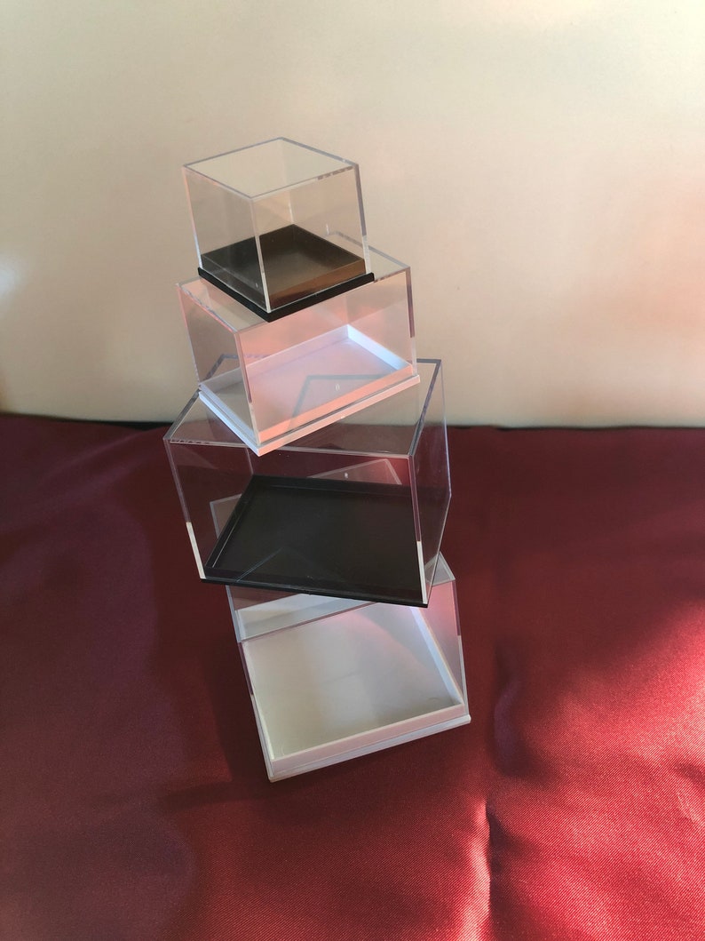 May include: Four clear acrylic display boxes stacked on top of each other. The top box has a brown base, the second box has a white base, the third box has a black base, and the bottom box has a white base.