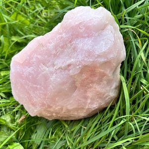 Può includere: Un grande pezzo grezzo di cristallo di quarzo rosa con un aspetto traslucido rosa pallido e una superficie irregolare e sfaccettata. La pietra presenta fratture naturali e vari livelli di opacità in tutta la sua struttura cristallina.