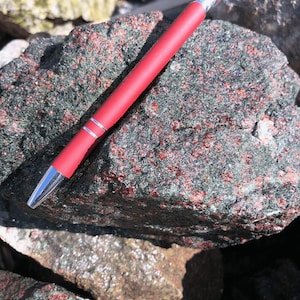 Peut inclure: Gros plan sur une roche vert foncé et rouge avec un stylo rouge sur le dessus. La roche a une texture rugueuse et est recouverte de petits cristaux rouges.