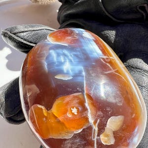 Puede incluir: Una piedra preciosa pulida, de forma ovalada, con una mezcla de colores. La piedra presenta una zona translúcida, azul claro y blanca, rodeada de tonos naranja y marrón. La superficie tiene un acabado brillante, con patrones e inclusiones naturales.