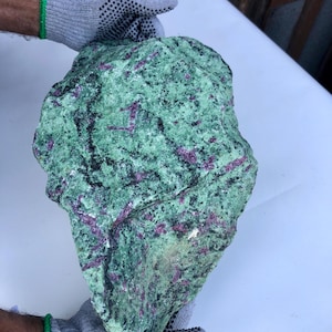 Könnte beinhalten: Ein großer, grob behauener Felsen mit einem gesprenkelten grün-roten Aussehen. Der Felsen wird von behandschuhten Händen gehalten, die seine Größe und Textur zeigen. Die Oberfläche des Steins ist eine Mischung aus grünen und roten Farbtönen mit einem natürlichen, unpolierten Aussehen.