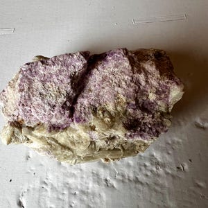 May include: A raw mineral specimen featuring a combination of vibrant purple crystalline sections and opaque, off-white to beige rocky material. The stone has a rough, uneven texture with visible jagged edges and natural mineral formations throughout.