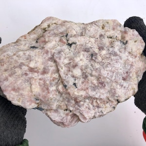 Peut inclure: Une grosse roche de forme irrégulière, avec un mélange d'inclusions minérales roses, blanches et noires. La surface de la roche a un aspect rugueux et texturé. La roche est tenue par des mains gantées.