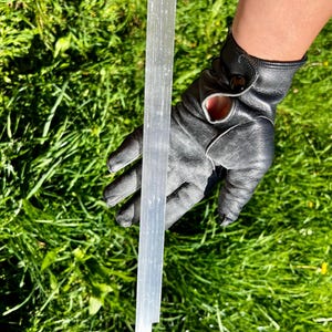 Könnte beinhalten: Ein langer, durchscheinender Selenit-Kristallstab, gehalten vor grünem Gras. Der Kristall hat eine blasse, cremefarbene Farbe mit sichtbaren Streifen. Der Stab ist etwa 25 cm lang.