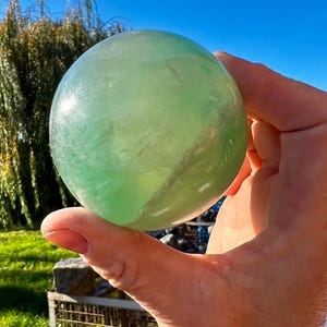 Könnte beinhalten: Eine transparente, grüne Fluoritkugel wird vor einem strahlend blauen Himmel gehalten. Der polierte Kristallball hat eine glatte Oberfläche und verschiedene Grüntöne. Die Kugel wird in einer Hand gehalten, mit einem verschwommenen Hintergrund aus Grün und einem Metallzaun.
