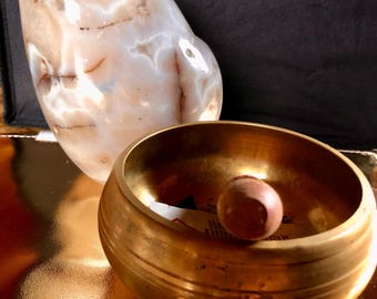 May include: A golden singing bowl with a wooden striker resting inside. The bowl is sitting on a gold-colored surface, next to a large, white agate stone.