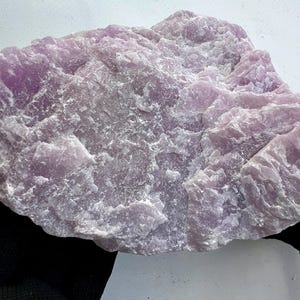 Könnte beinhalten: Ein großes, grob behauenes Stück Mineral in Lila. Der Stein hat eine strukturierte, unebene Oberfläche mit verschiedenen Lila- und Weißtönen. Das Mineral scheint ein natürliches, unpoliertes Exemplar zu sein.