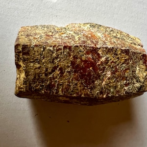 May include: A raw, unpolished mineral specimen with a rough, textured surface. The stone displays a variegated color palette of deep amber, burnt orange, and earthy brown tones, with visible crystalline inclusions and a jagged, irregular shape.