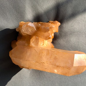 Puede incluir: Un grupo de cristales de cuarzo de color naranja claro. El cristal tiene una base grande y rectangular con un grupo de cristales más pequeños en la parte superior. La superficie tiene una textura rugosa y es translúcida.