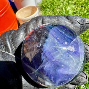 Könnte beinhalten: Eine transparente Kristallkugel mit inneren blauen und violetten Adern. Die Kugel wird in einem schwarzen Lederhandschuh gehalten. Der Hintergrund zeigt Eimer und grünes Gras.