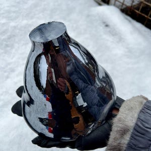May include: A dark grey, reflective glass vase with a rounded body and a flat, angled top. The vase is held in a gloved hand against a snowy backdrop. The surface of the vase reflects the surroundings.