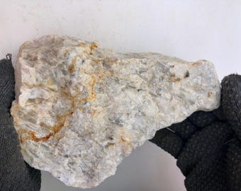 May include: A large, irregularly shaped rock with a rough, textured surface. The rock is primarily off-white with streaks of tan and gray. The rock is held by a person wearing a black glove.