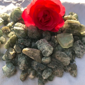 May include: A red rose sits on a pile of green and gray crystals. The crystals are scattered on a white surface.