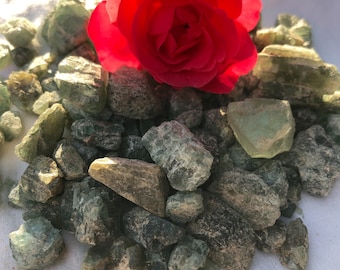 May include: A red rose sits on a pile of green and gray crystals. The crystals are scattered on a white surface.