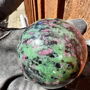 Op de afbeelding: Een gepolijste edelsteenbol met een mix van groene, roze en zwarte kleuren. De bol wordt vastgehouden in een zwarte leren handschoen. De edelsteen heeft een gespikkeld patroon.
