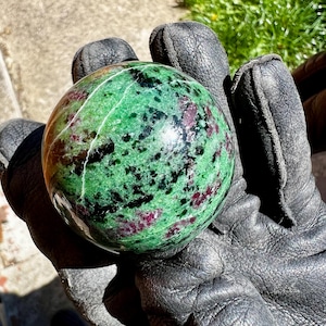 Può includere: Una sfera lucida di zoisite verde e rossa, tenuta in una mano guantata. La pietra presenta un motivo maculato di tonalità verde scuro, nero e rosso intenso. La superficie della sfera è liscia e riflettente.