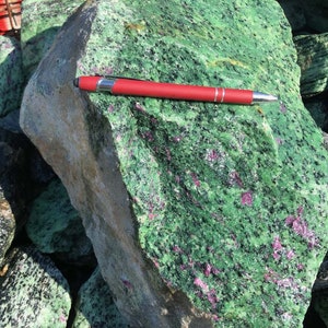 May include: A collection of large, rough-cut green zoisite stones with ruby inclusions. A red pen is placed on one stone for scale. The stones display a speckled pattern with red and black flecks within the green matrix.