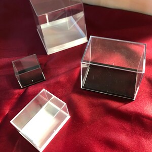 May include: Four clear acrylic display boxes of varying sizes. The boxes have white bases and black felt lining. The boxes are arranged on a red surface.