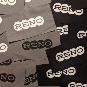 Könnte beinhalten: Nahaufnahme von mehreren Stoff-Patches mit dem Wort "RENO" in weiß auf schwarzem oder grauem Hintergrund. Die Patches sind in einem zufälligen Muster angeordnet.