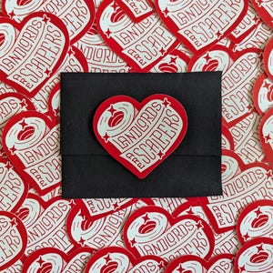 May include: A collection of red heart-shaped stickers with the text "Landlords Are Scapers" in white. One sticker is placed on a black envelope. The stickers have a retro design with stars and a coffee bean graphic.