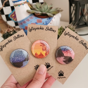 Op de afbeelding: Drie ronde knopen met verschillende ontwerpen op bruin karton. De knopen zijn alle 2,5 cm in diameter. De eerste knop heeft een blauw en wit sterrenhemelontwerp. De tweede knop heeft een rood, oranje en geel zonsondergangontwerp. De derde knop heeft een roze en blauw bergontwerp. Elke kaart heeft de tekst "Lytsepoekie Buttons" en de social media handle "@lytsepoekie".