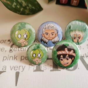 Op de afbeelding: Vijf ronde groene en witte knoppen met cartoonfiguren. De figuren zijn een groen wezen met een wit masker, een man met wit haar en een baard, een groen wezen met een groene mohawk en twee mensen met zwart haar.