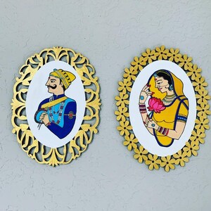 May include: Two oval-shaped wall art pieces. One features a man in a blue and gold outfit, the other a woman in yellow and gold, both with traditional Indian attire. The frames are gold-colored with intricate designs and floral patterns.