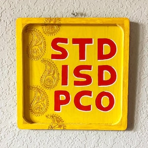 May include: A square, yellow wooden sign with the letters "STD ISD PCO" in red, outlined in white. The sign has a decorative paisley pattern in the corners.