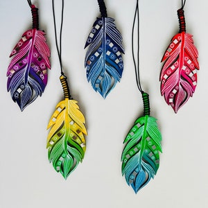 May include: A collection of six feather-shaped pendants in various colors, including purple, blue, red, yellow, and green. Each pendant has a black cord and a decorative top. The feathers have a mirrored center and white detailing.