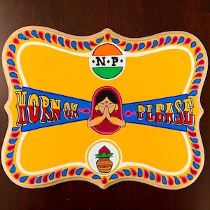 May include: A hand-painted wooden sign with a yellow background and a decorative border. The sign features the text "HORN OK PLEASE" in red and blue, along with an illustration of a woman with folded hands and other Indian cultural elements.