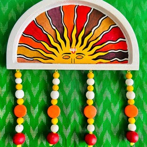 May include: A white, semi-circular wall hanging with a painted sun design in shades of red, orange, yellow, and brown. Below, four strands of beads in white, yellow, orange, and red hang from the sun. The background is a green fabric with a chevron pattern.