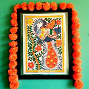 May include: A framed artwork featuring a colorful peacock illustration with orange, yellow, and blue hues. The artwork is surrounded by a black frame and adorned with a garland of orange pom-poms, set against a green wall. The text "@DoDevyan" is visible.