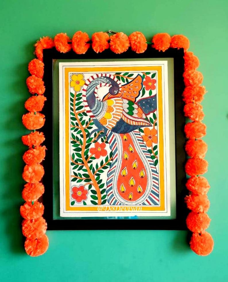 May include: A framed artwork featuring a colorful peacock illustration with orange, yellow, and blue hues. The artwork is surrounded by a black frame and adorned with a garland of orange pom-poms, set against a green wall. The text "@DoDevyan" is visible.