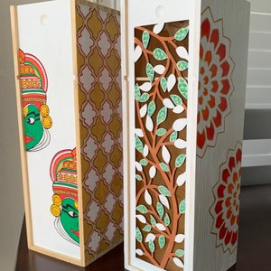 May include: Two wooden boxes with decorative designs. One box features a traditional Indian face painting, the other has a cut-out tree design. Both have white and gold accents and rope handles.