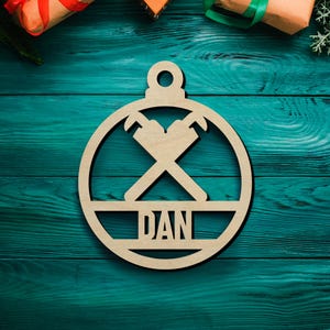 May include: A circular light-colored wooden ornament featuring a cutout design of two crossed pipe wrenches in the upper section and the name DAN in capital letters within a horizontal bar across the center, topped with a small hanging loop.