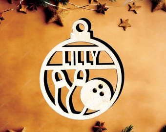 May include: A light-colored wooden Christmas ornament shaped like a sphere with a cutout design featuring two bowling pins and a bowling ball. The name LILLY is cut out in the upper section of the ornament, which includes a circular hanging loop.