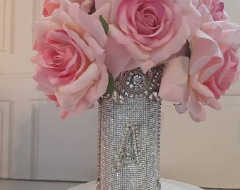 May include: A silver vase covered in rhinestones with a pink rose bouquet. The vase has a decorative design at the bottom. The vase is sitting on a white surface.