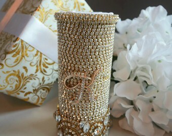 May include: A gold-colored vase covered in rhinestones with a large rhinestone letter "R" on the front. The vase is sitting on a white surface next to a white gift box with gold accents and a white bouquet of flowers.