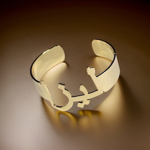 Puede incluir: Una pulsera de metal de color dorado con un diseño recortado de escritura árabe.