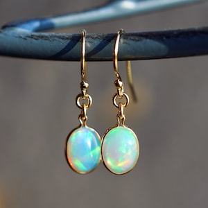 Könnte beinhalten: Ein Paar goldene Ohrhänger mit schillernden Opal-Edelsteinen. Die Ohrringe haben ein schlichtes Design mit einem runden Opal-Stein, der an einer goldenen Öse hängt.