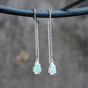 May include: Silver threader earrings featuring pear-shaped opal pendants. The opals display a milky white base with flashes of blue and green. The earrings are suspended from fine silver chains.