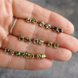 May include: A collection of various green gemstone stud earrings in different shapes and sizes. The earrings are set in silver and displayed on a hand. The gemstones range in color from light to dark green.