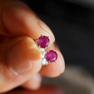 Peut inclure: Une paire de boucles d'oreilles à clous en rubis. Chaque boucle d'oreille présente une pierre précieuse rouge vif et ronde, sertie dans un sertissage à griffes en argent ou en métal blanc. Les boucles d'oreilles sont présentées sur fond sombre.